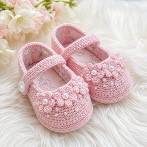 May include: A pair of pink crochet baby shoes. Each shoe has a strap with a button fastening and is decorated with a flower design and small pearl accents. The interior lining has a floral pattern.