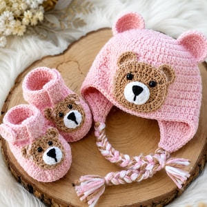 May include: A pink crochet baby set featuring a hat with bear ears and a bear face, and matching booties. The hat has a braided tie with pink and white tassels. The booties and hat have brown bear faces with black eyes and white snouts.