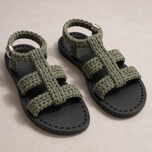 May include: A pair of sandals featuring olive green crocheted cord straps in a gladiator style. The sandals have a black textured footbed with a perforated edge and a silver-toned metal buckle on the ankle strap for a secure fit.