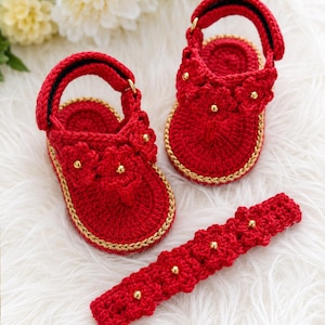 Puede incluir: Un conjunto de sandalias y diadema de ganchillo rojas para bebé. Las sandalias tienen ribetes dorados y pequeños detalles florales. La diadema presenta detalles florales similares y cuentas doradas. El conjunto se muestra sobre una superficie blanca y esponjosa.