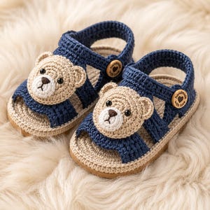 Op de afbeelding: Een paar handgemaakte babysandalen. De sandalen zijn gehaakt in marineblauw en beige. Elke sandaal heeft een teddybeergezicht en een houten knoopsluiting. De sandalen liggen op een zacht, wit oppervlak. De sandalen zijn ongeveer 10 cm lang.