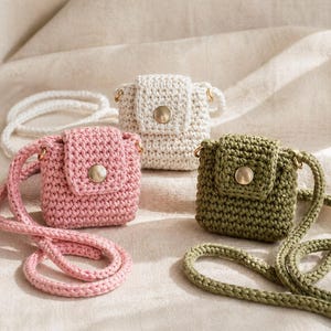 Può includere: Tre piccole borse a tracolla all'uncinetto in rosa, bianco e verde oliva. Ogni borsa presenta una chiusura con patta con un bottone color oro e un cinturino abbinato. Le borse sono esposte su un tessuto dai toni neutri.