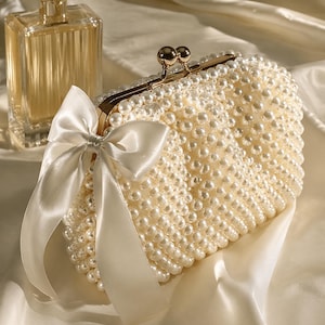 May include: Ivory pearl-covered clutch with a gold clasp and a white satin bow. A rectangular perfume bottle with a gold cap sits nearby. The clutch is a classic accessory for special occasions.