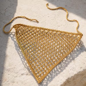 Puede incluir: Un pañuelo triangular de ganchillo dorado con un patrón de red. El pañuelo tiene un tejido delicado y abierto y largas cintas trenzadas en las esquinas superiores. Está sobre una superficie clara y texturizada.