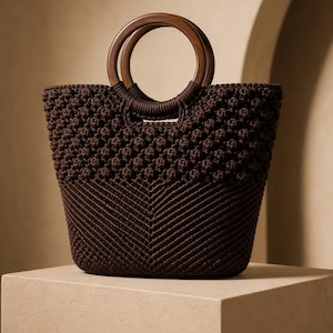 May include: A brown macrame tote bag with wooden handles. The bag features a textured, knotted design and a chevron pattern on the lower half. The circular wooden handles provide a contrasting element to the woven body.