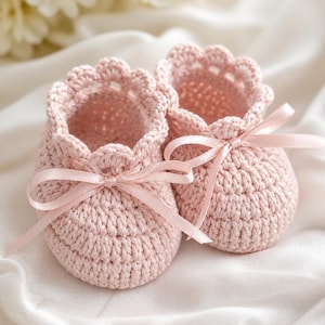 Op de afbeelding: Een paar handgemaakte, lichtroze gehaakte babyslofjes. Elk slofje heeft een geschulpte rand en is vastgebonden met een bijpassend roze satijnen lint. Het zachte, getextureerde garen creëert een delicate en gezellige uitstraling.
