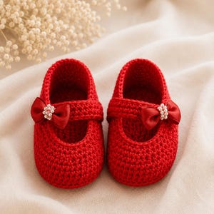 May include: A pair of bright red, crocheted baby shoes. Each shoe features a strap with a red satin bow adorned with small, pearl-like beads. The shoes are designed with a rounded toe and a delicate, handmade appearance.