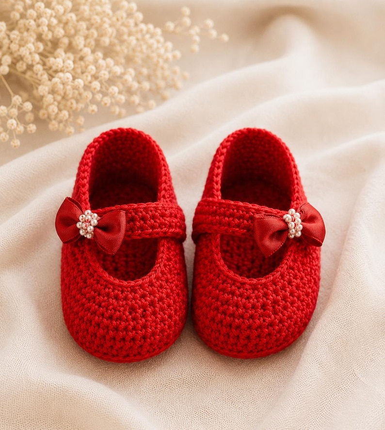 May include: A pair of bright red, crocheted baby shoes. Each shoe features a strap with a red satin bow adorned with small, pearl-like beads. The shoes are designed with a rounded toe and a delicate, handmade appearance.