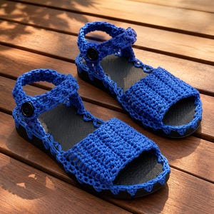 Puede incluir: Un par de sandalias azules de ganchillo hechas a mano. Las sandalias presentan un diseño de ganchillo texturizado con una correa que se abrocha con un botón negro. Las suelas son negras. Las sandalias se exhiben sobre una superficie de madera.