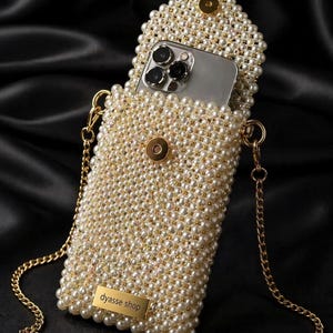 Puede incluir: Bolso para teléfono con cuentas de perlas color marfil con una correa de cadena dorada. La bolsa está abierta, revelando un teléfono inteligente en el interior. La bolsa tiene un cierre dorado y una pequeña etiqueta rectangular dorada que dice "dyasse shop".