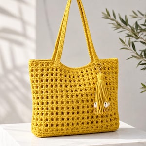 May include: A mustard yellow crochet tote bag featuring an open-weave grid pattern, two long shoulder straps, and a matching yarn tassel attached to a gold-tone ring. The tassel is decorated with small white cowrie shells at the bottom.