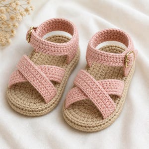 Pode incluir: Um par de sandálias de bebê de crochê feitas à mão. As sandálias apresentam tiras cruzadas rosa claro e uma tira no tornozelo com fivela dourada. As solas são bege natural. Um acessório charmoso para bebês.