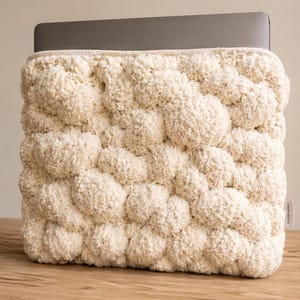 Può includere: Custodia per laptop color crema con un design strutturato e a bolle. La custodia è realizzata in un materiale morbido e felpato ed è progettata per proteggere un laptop. Il laptop è parzialmente visibile nella parte superiore. La custodia è appoggiata su una superficie di legno.