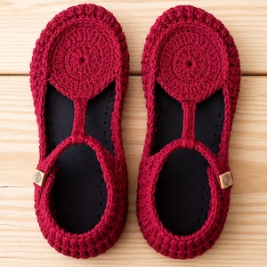 May include: A pair of deep red crochet slippers featuring a circular pattern on the top, a T-strap design, and a black inner sole. Each slipper includes a small square wooden tag on the side with a circular spiral logo engraved on the surface.