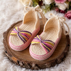 Op de afbeelding: Een paar handgemaakte gehaakte sandalen in een neutrale beige kleur met roze en veelkleurige accenten. De sandalen hebben een strikdetail aan de voorkant en een bandje dat bij de enkel wordt vastgemaakt. Ze worden gepresenteerd op een houten schijf.