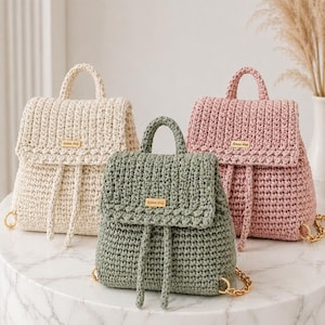 May include: Three crochet backpacks in cream, sage green, and pink. Each bag has a flap closure, a top handle, and a drawstring. Gold-colored chain straps and a small gold tag with the text "dyasse shop" are visible on each bag.