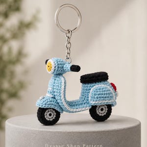 May include: A light blue crocheted scooter keyring featuring a black seat, black wheels with grey centres, a yellow headlight, and a small red taillight. The scooter is attached to a silver metal split ring and chain. Text reads: Dyasse Shop Pattern.
