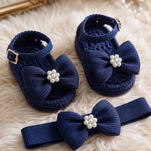 Puede incluir: Conjunto de sandalias y diadema de bebé de crochet azul marino. Las sandalias tienen un lazo con detalles de perlas y correas ajustables en el tobillo con hebillas doradas. La diadema a juego también tiene un lazo con adornos de perlas.