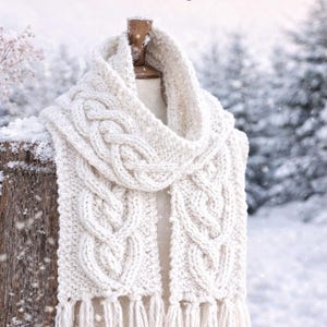 Könnte beinhalten: Cremefarbener Zopfstrickschal mit Herzmuster, präsentiert auf einem Holzständer vor verschneitem Hintergrund. Der Schal hat Fransen am unteren Ende. Der Text auf dem Bild lautet "Heart Cable Scarf" und "Dyasse Designs."
