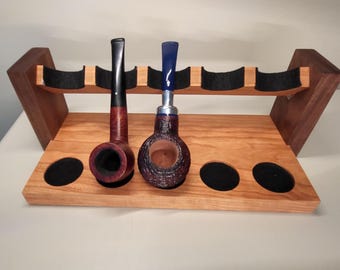 May include: Wooden pipe rack with two pipes. The rack is made of light brown wood with a dark brown accent. The pipes are dark brown and blue. The rack has black felt supports for the pipes.