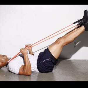 May include: A person lying on their back with their legs raised and secured with a red resistance band. The person is holding the band with both hands and is pulling it towards their chest.