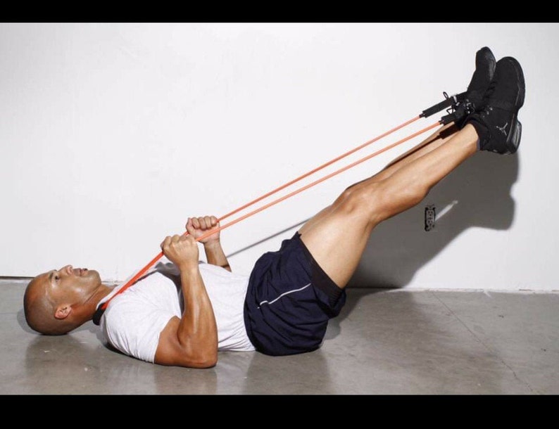 May include: A person lying on their back with their legs raised and secured with a red resistance band. The person is holding the band with both hands and is pulling it towards their chest.