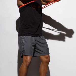 May include: A man wearing a black tank top and gray shorts is using a resistance band for a workout. He is standing on a white surface and wearing black sneakers.