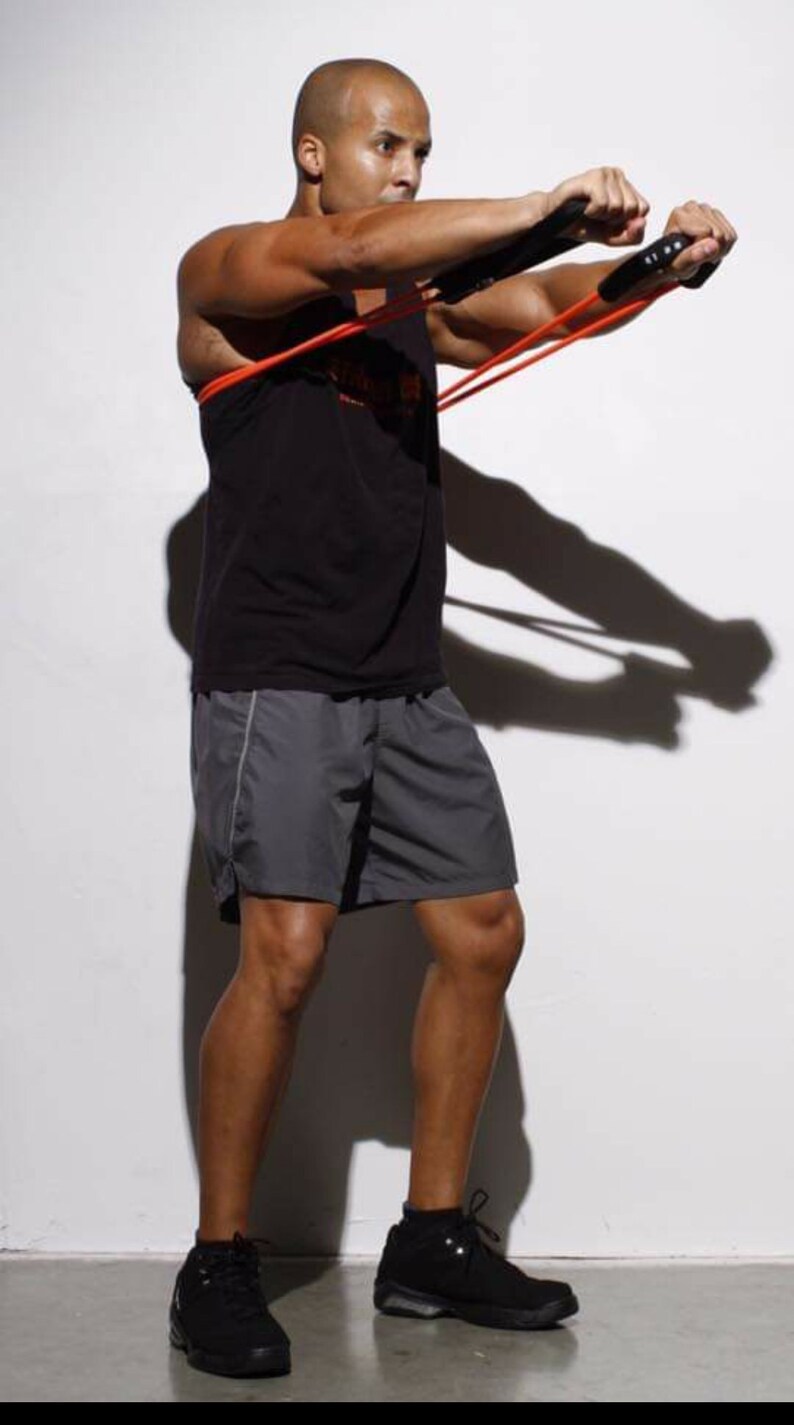 May include: A man wearing a black tank top and gray shorts is using a resistance band for a workout. He is standing on a white surface and wearing black sneakers.