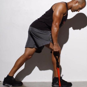 May include: A man wearing black shorts and a black tank top is using a red resistance band to do a leg exercise. He is standing with his left leg straight and his right leg bent at the knee.