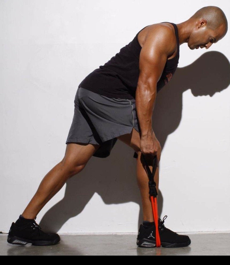 May include: A man wearing black shorts and a black tank top is using a red resistance band to do a leg exercise. He is standing with his left leg straight and his right leg bent at the knee.