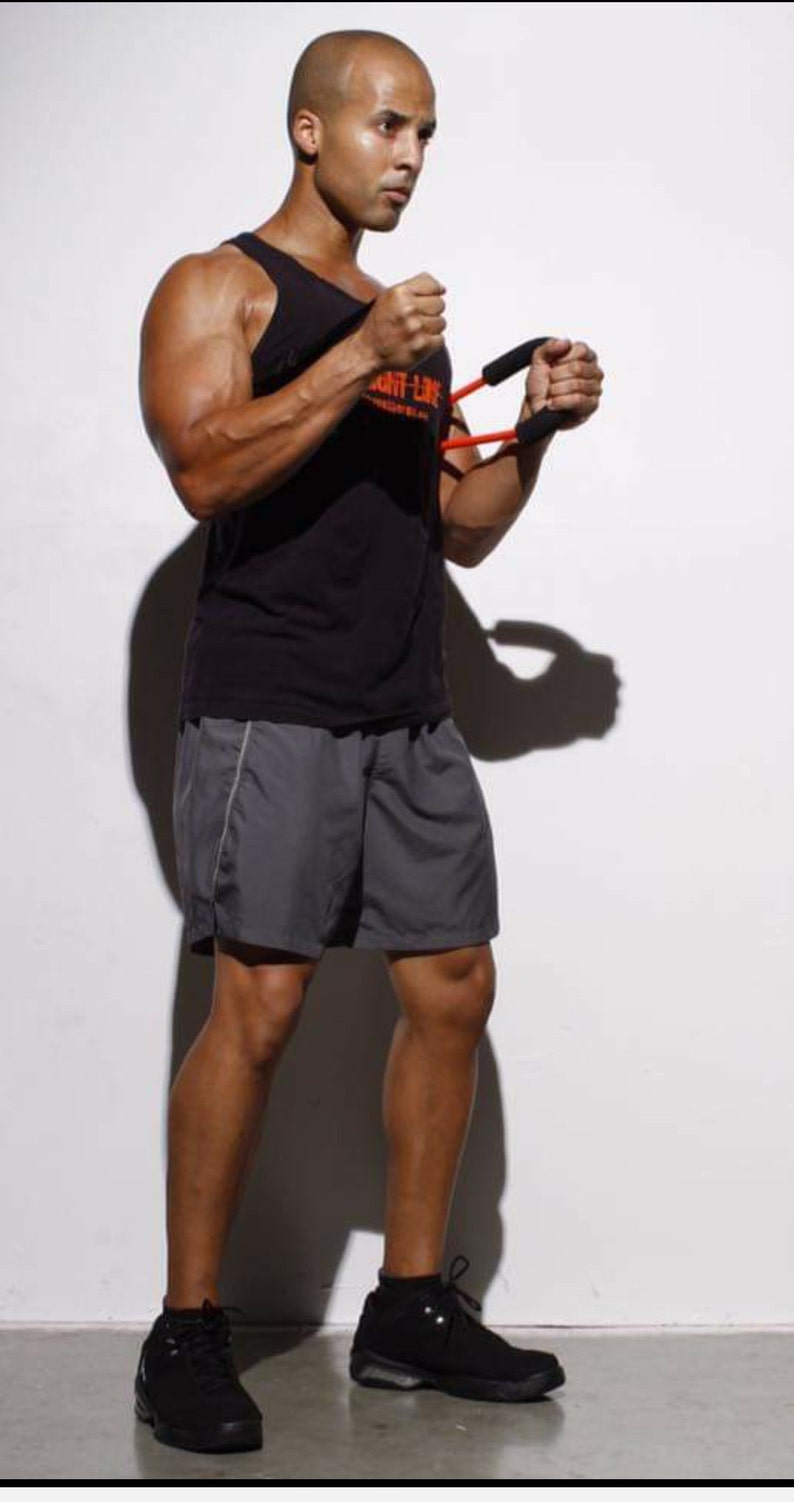 May include: A man wearing a black tank top with the text "Fight Like" on it, gray shorts, and black sneakers. He is holding a red and black resistance band in his hands.