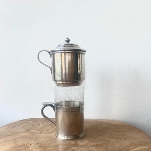 May include: Silver coffee maker with a glass filter and a silver cup. The coffee maker has a lid with a knob on top.