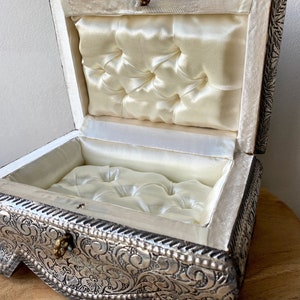 May include: A silver metal jewelry box with an ornate floral design. The box is open and lined with white satin.