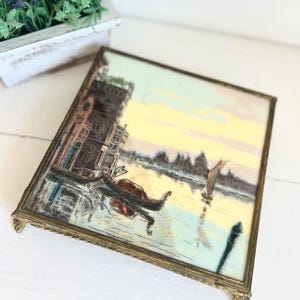 May include: A vintage square trinket tray with a detailed painting of a Venetian canal scene. The artwork features gondolas, buildings, and a sunset. The tray has a decorative gold-toned metal frame and feet.