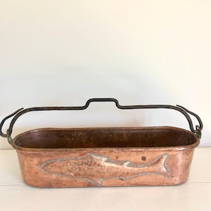 Peut inclure: Une poissonnière en cuivre de forme oblongue avec une patine vieillie et une anse pivotante en métal sombre. L'extérieur présente un motif de poisson en relief sur le côté, mettant en valeur des écailles et des nageoires détaillées.