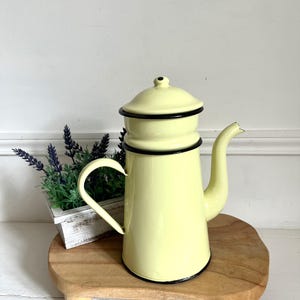 Op de afbeelding: Een lichtgele emaille koffiekan met een zwarte rand en handvat. De kan heeft een ingebouwd filter en staat op een houten standaard.