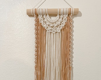 May include: A macrame wall hanging with a wooden dowel at the top. The macrame is made with white and beige yarn and has a fringe at the bottom.
