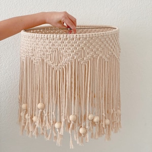 May include: A beige macrame lampshade with wooden beads hanging from the bottom. The lampshade is made of a thick, natural fiber rope and has a fringe design.