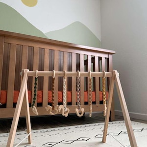 May include: A wooden baby play gym with hanging toys in various colors and shapes. The gym is set up on a patterned play mat in front of a wooden crib. The background features a wall with a painted mountain scene and a yellow sun.