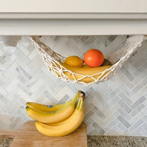 May include: A white string hammock hanging from a white cabinet, holding a bunch of bananas and an orange. A wooden stand with four bananas on top sits on a countertop with a white and gray tiled backsplash.