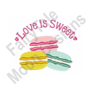 May include: Three colorful macarons with the text "Love is Sweet" in pink lettering and pink hearts on either side of the text.