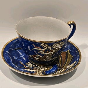 May include: A blue and white porcelain teacup and saucer set. The cup and saucer feature a detailed dragon design with gold accents. The rim of the cup and saucer are trimmed with gold. A classic tea set.