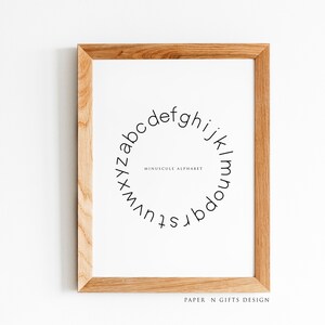 May include: A framed print with a white background and a black circle containing the lowercase alphabet in a circular pattern. The text "MINUSCULAR ALPHABET" is centered inside the circle. The frame is made of light brown wood. The text "PAPER N GIFTS DESIGN" is printed at the bottom of the print.