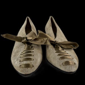 May include: A pair of beige suede shoes with pointed toes and olive green satin ribbon laces. The shoes are set against a black background, highlighting their vintage style and design.