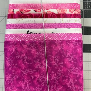 May include: A stack of pink fabric squares tied with silver string. The fabrics feature various patterns, including butterflies, polka dots, floral designs, and swirls. The color palette ranges from light pink to deep magenta, creating a visually appealing bundle.