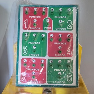 Op de afbeelding: Een rechthoekig spelbord met groene en rode secties, elk met nummers en het woord "PUNTOS". Het bord is in plastic verpakt. De nummers 0-9 worden weergegeven. De tekst "CHICOS" is ook zichtbaar.