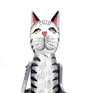 May include: A wooden cat figurine with gray and black stripes, painted with yellow eyes, a pink nose, and red ears. The cat is sitting on a blue and white base with its arms and legs outstretched.
