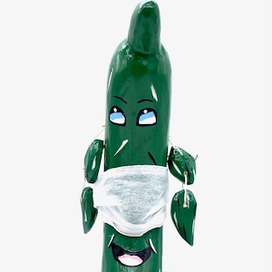 May include: A green wooden puppet shaped like a cucumber wearing a white face mask. The puppet has a painted face with blue eyes and a pink mouth. The puppet is sitting on a blue glass base.