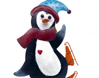 May include: A black and white penguin wearing a blue and white hat and a red scarf is ice skating. The penguin is holding a red heart. The ornament is decorated with blue and white beads and a blue heart.