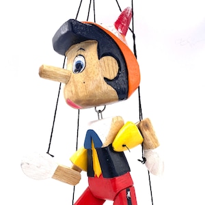 May include: A wooden Pinocchio puppet with a long nose, painted in bright colors, including red, yellow, blue, and black. The puppet is suspended by strings and has movable arms and legs.
